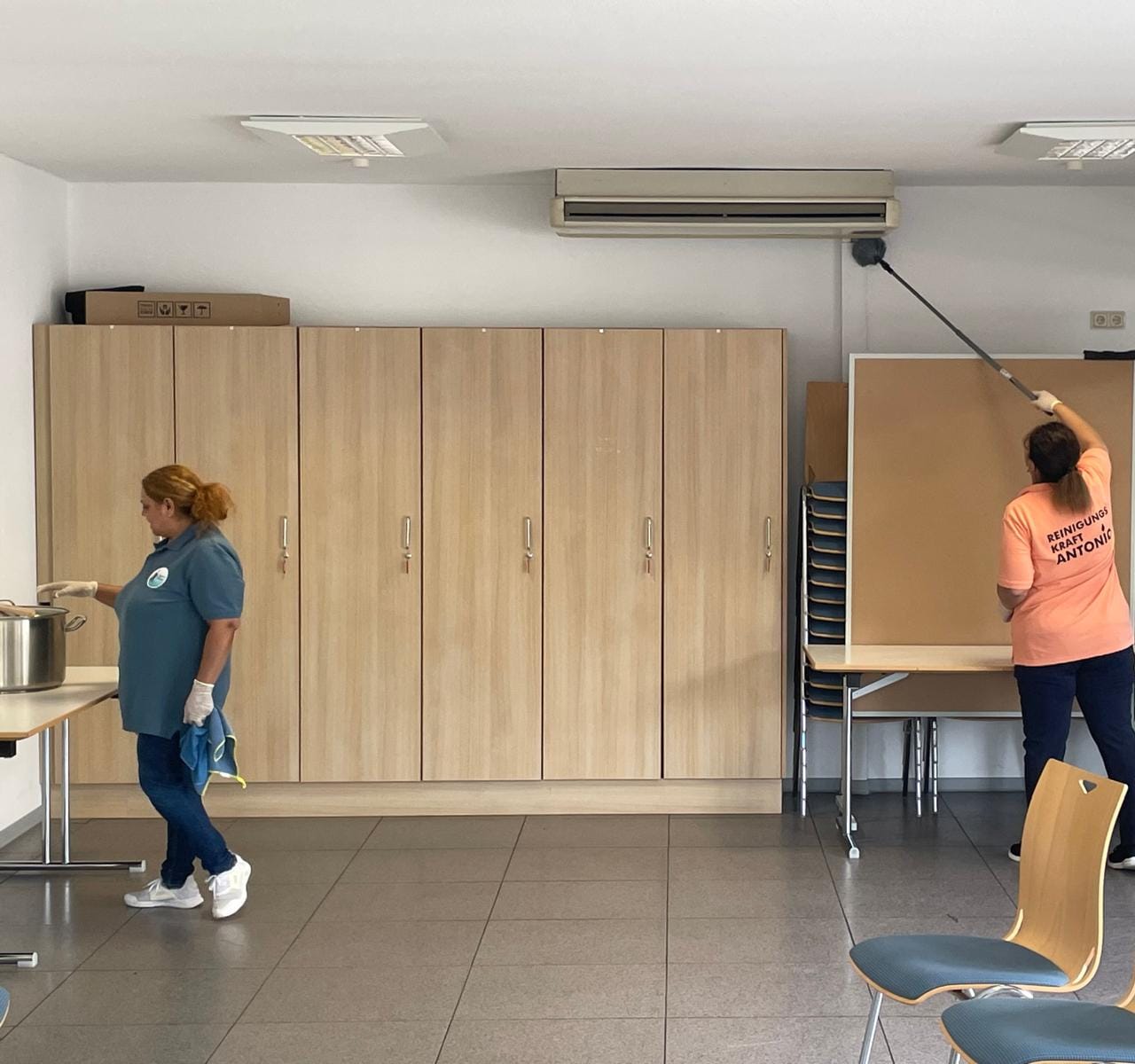 Zwei Mitarbeiterinnen reinigen einen Gemeinschaftsraum, Staubwedel an der Wand